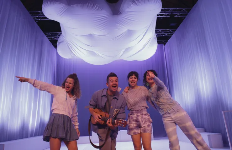 cuatro personas en un escenario con telas blancas, sonriendo y señalando con los brazos extendidos