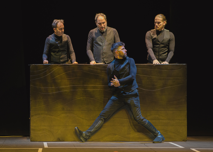 cuatro hombres en un escenario tres en un plano superior con una gran caja delante el cuarto delante con gesto de estar andando